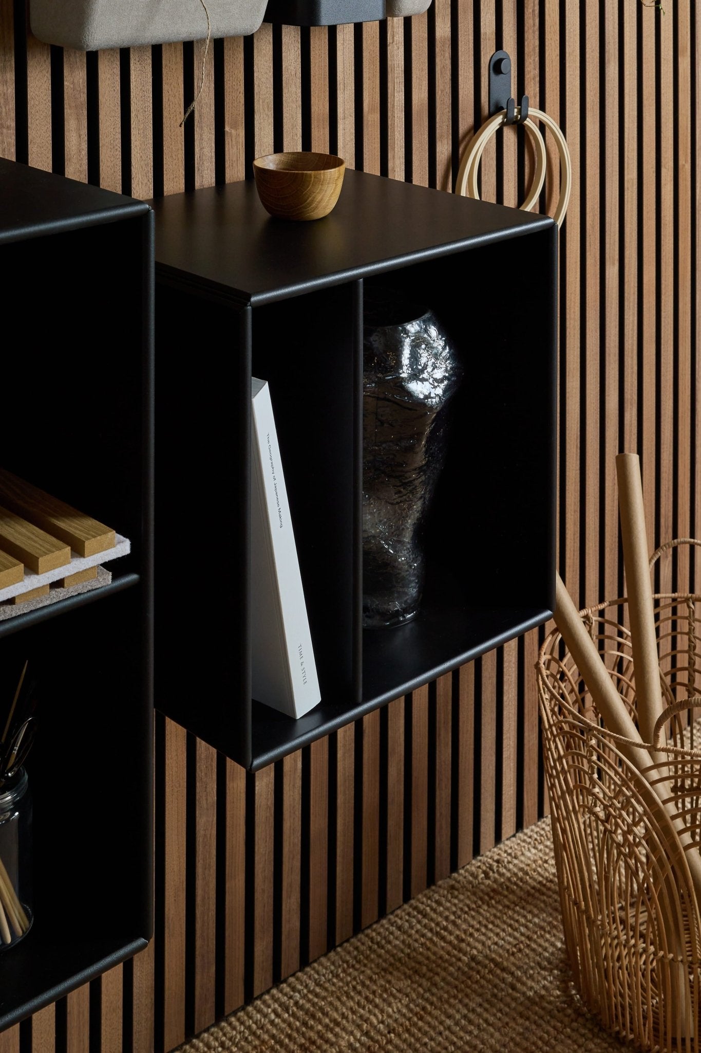 The Arka Bookcase / Black by WoodUpp, a modular shelf with two cubbies, placed beside a wicker basket and wooden bowl on a Nordic acoustic slatwall panel.